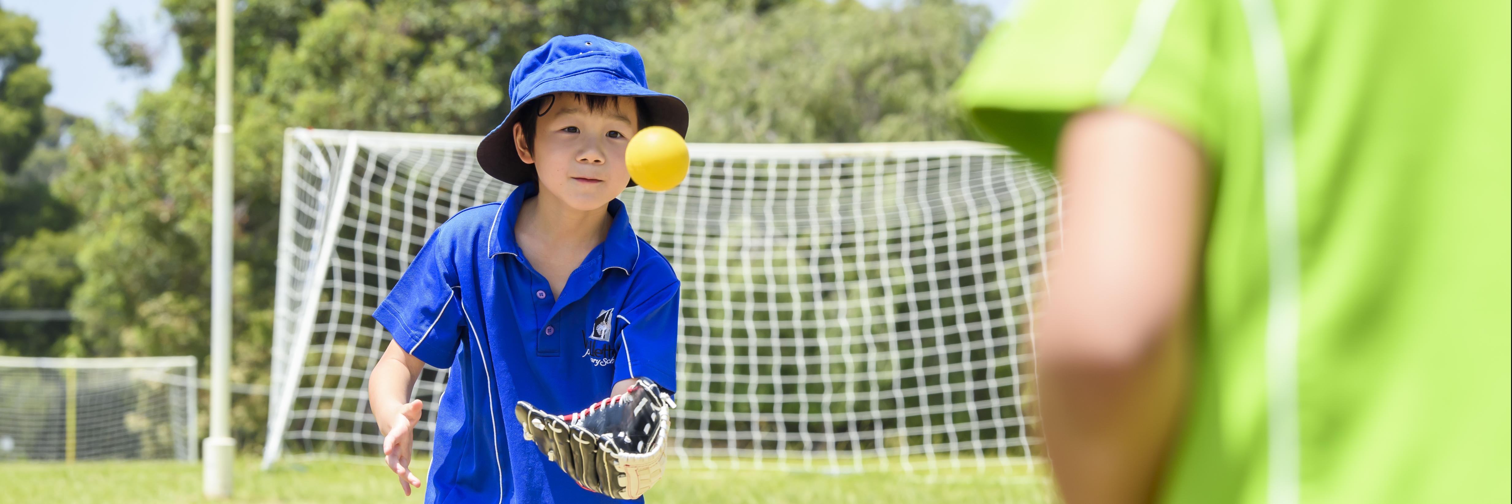 boy with a blue faction shirt and hat on looks towards a yellow tee ball as he tries to catch it in his glove. to the right of image is a boy with a green faction shirt on who had thrown him the ball