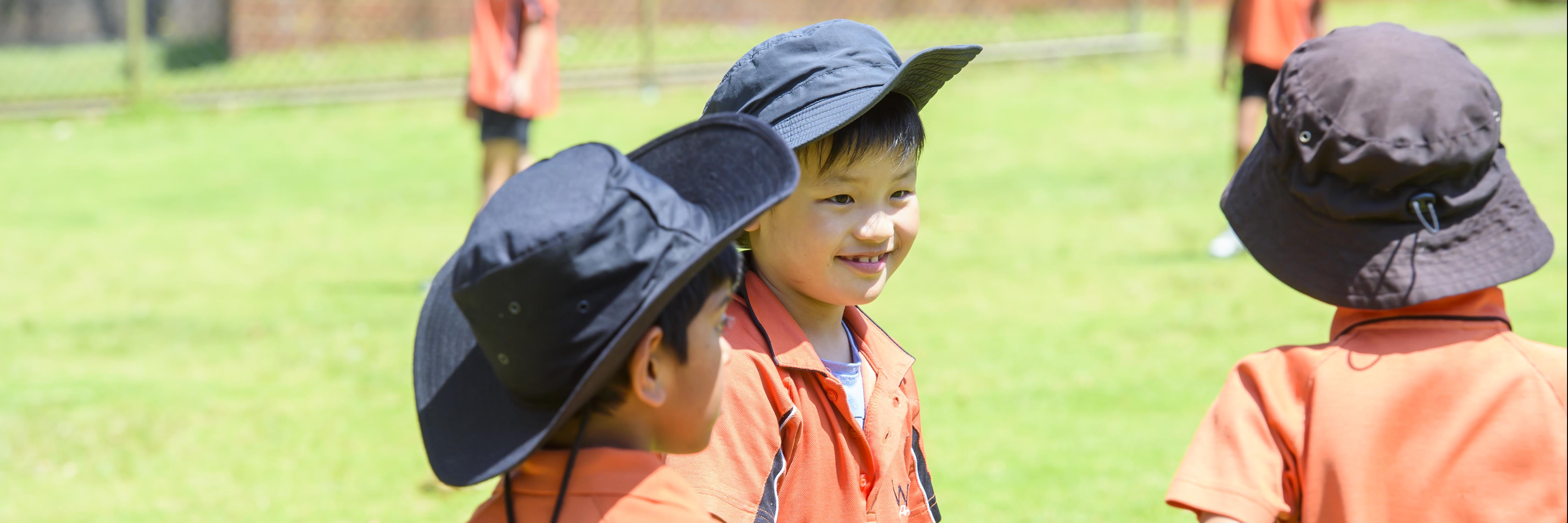 Three boys wearing hats smiling at each other during sport on the oval