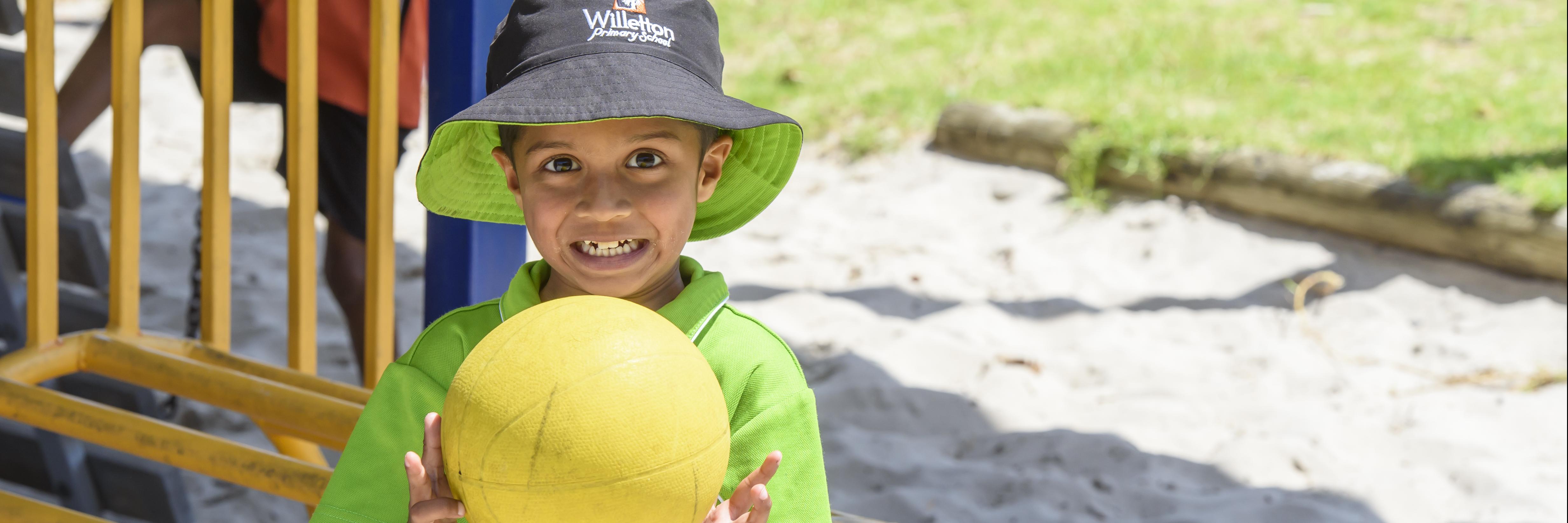 boy with green shirt and hat on holding a yellow soccer ball to his chest, smiling at camera. Playground in background of image