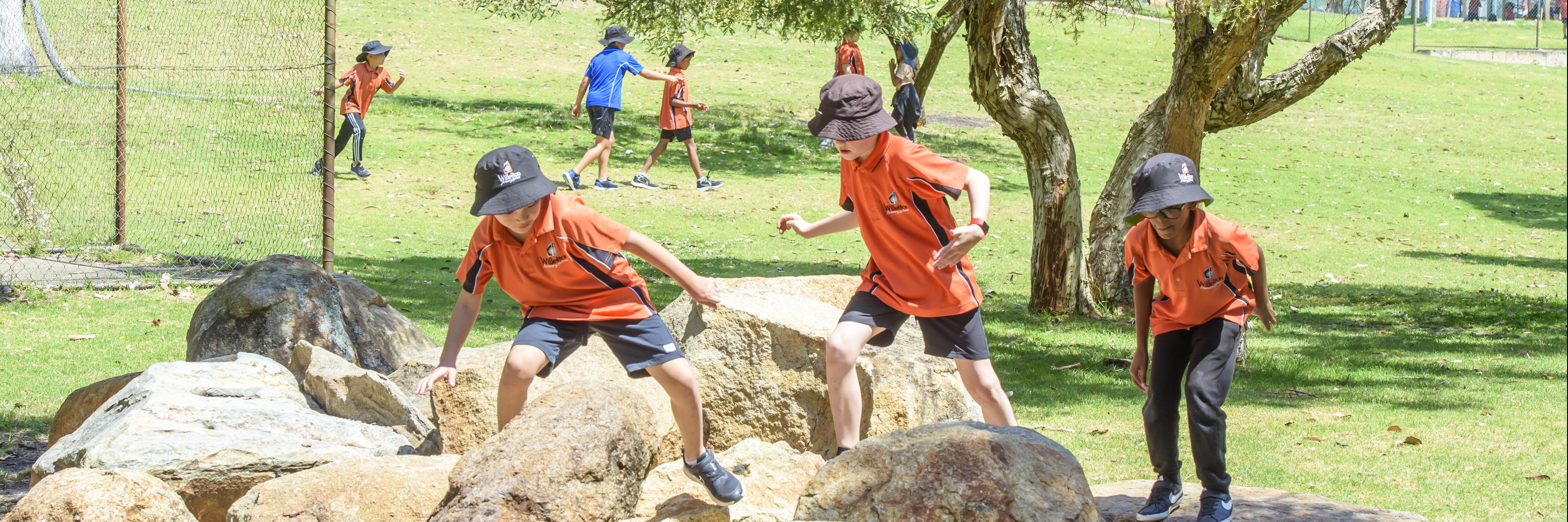 3 boys taking in turns climbing and jumping over large yellow rocks in the playground, all boys looking down towards their feet and holding onto various rocks