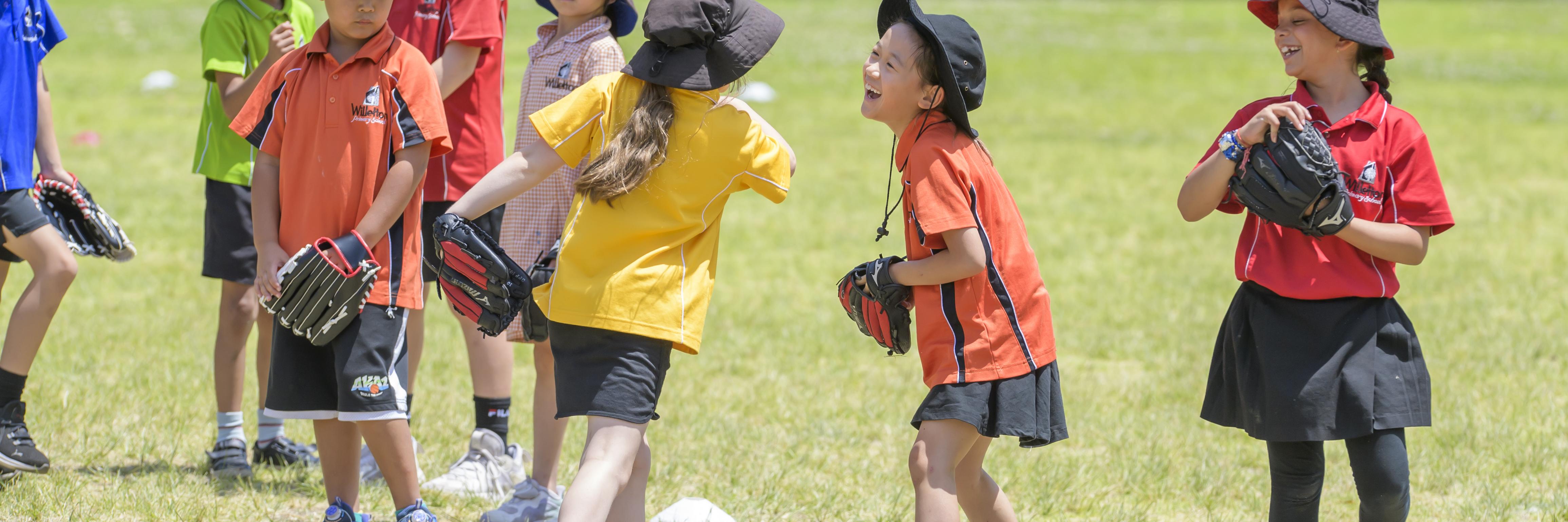 girl 1 is in a yellow faction shirt and has a tee ball glove on. she is walking away from the camera towards girl 2 who is facing towards camera and is laughing at girl 1. girl 3 is in a red faction shirt and is off center to the right, looking and smiling towards girl 1 and 2 in center frame.