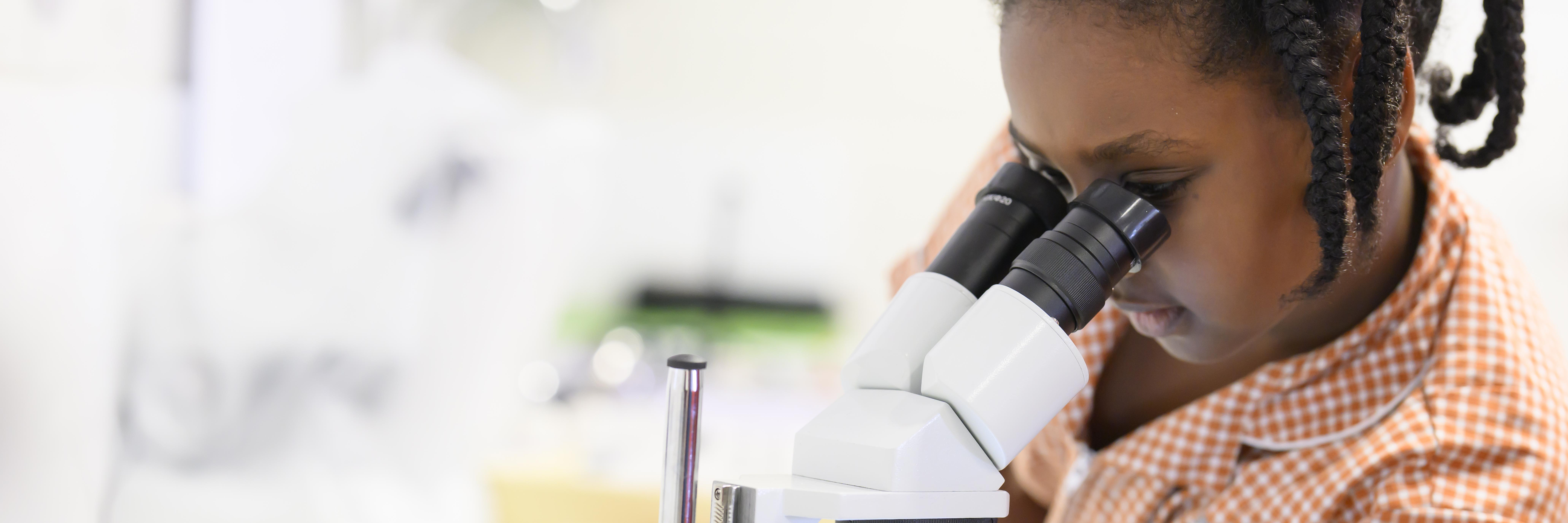 girl on right hand side of image looking into a white microscope with black eyepieces to her left. 