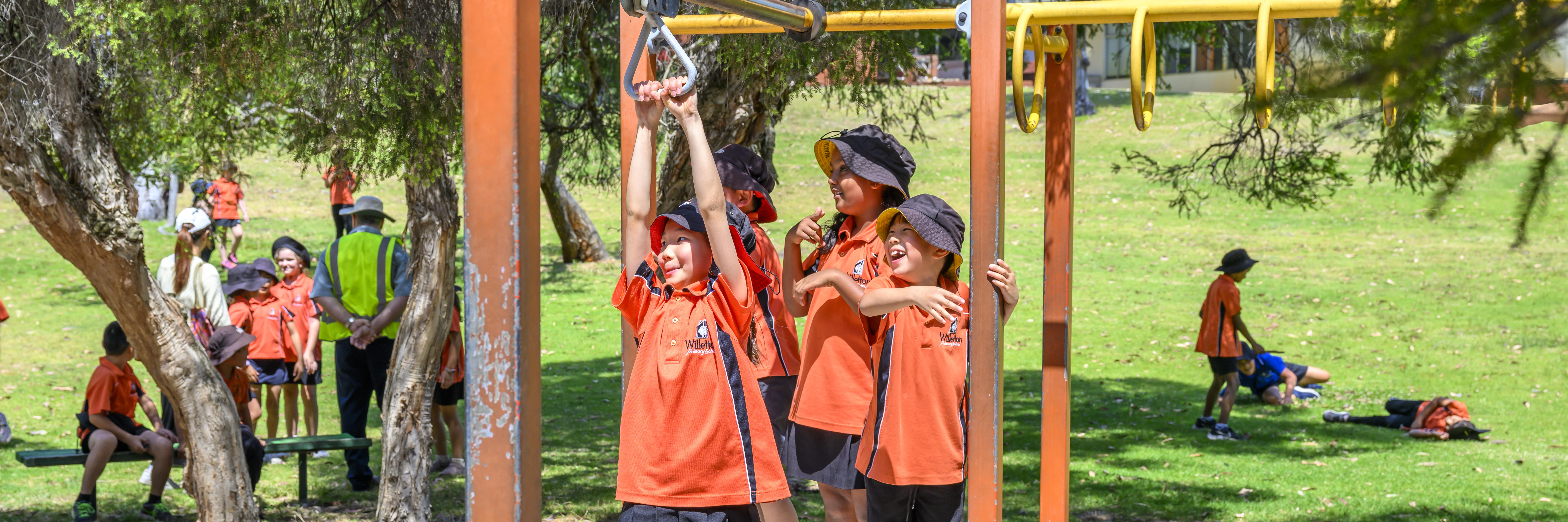 children out in the playground in background of image. front of image is a girl riding on a flying fox, looking left of camera as she holds onto the handles above her. children waiting in line behind her are smiling and laughing, watching her cross the flying fox.