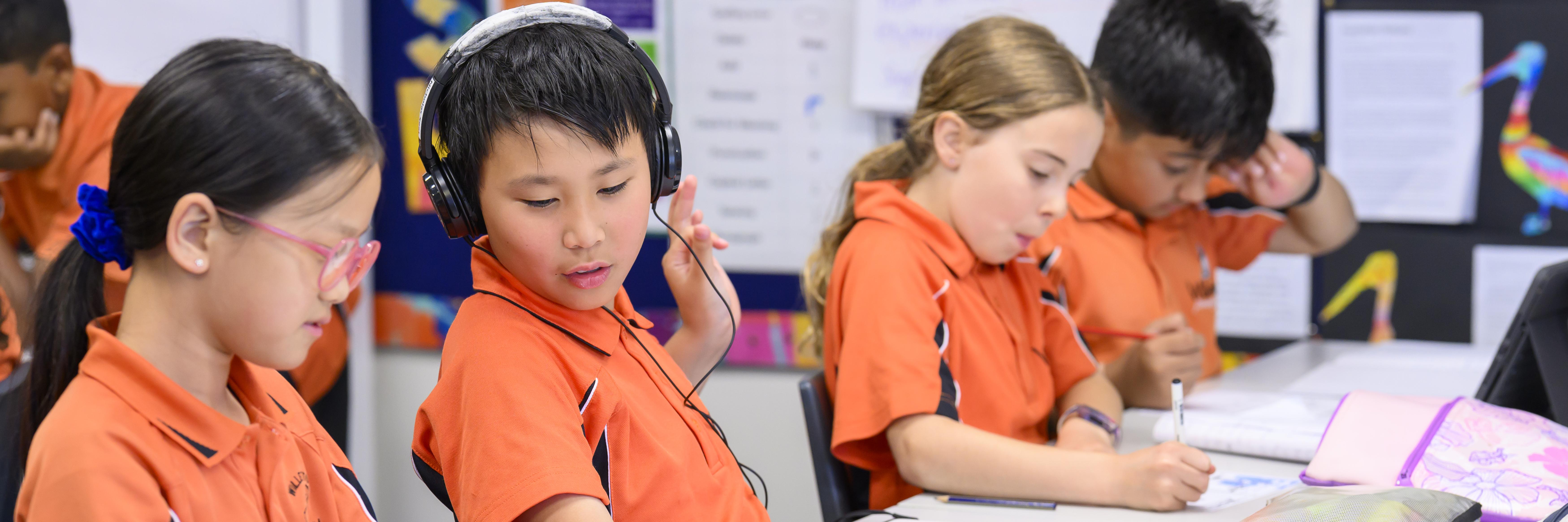 boy 1 with headphones on in center of image looking down towards Girl 1's work, talking to her. girl 2 and boy 2 in back of image out of focus, writing in their workbooks